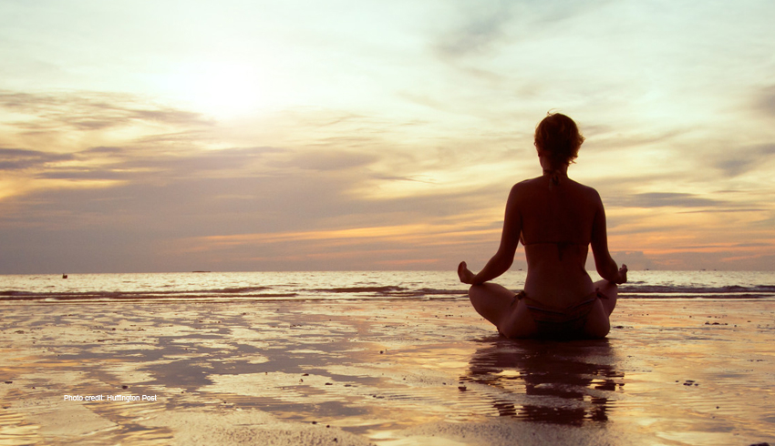 A woman in a bathing suits sits and mediates on the beach. 